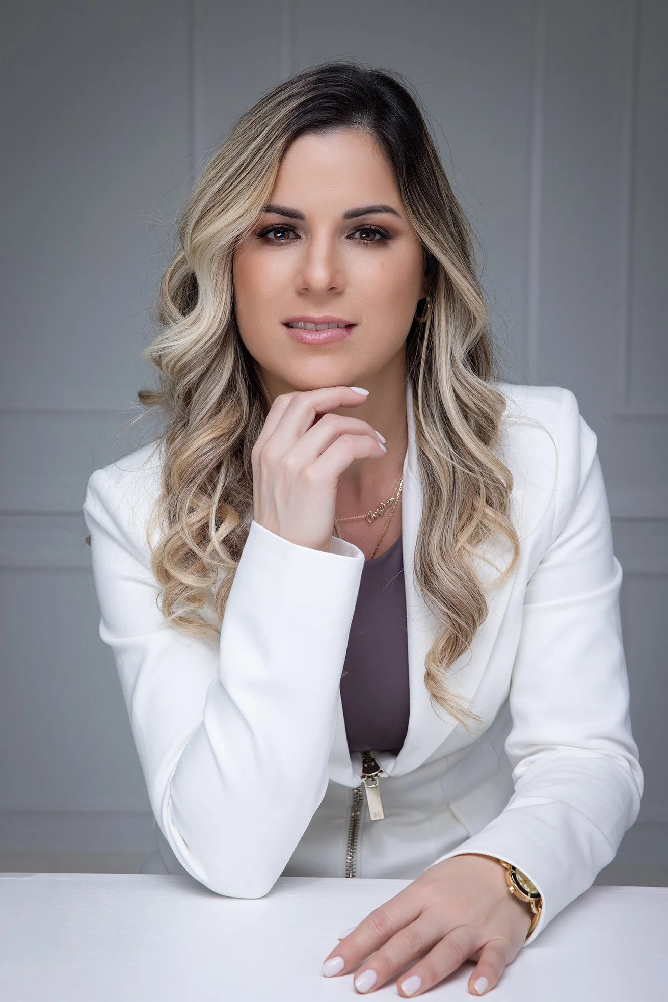A woman with styled blonde hair and makeup, wearing a white blazer, gold jewelry, and a watch, sitting at a desk and looking at the camera.