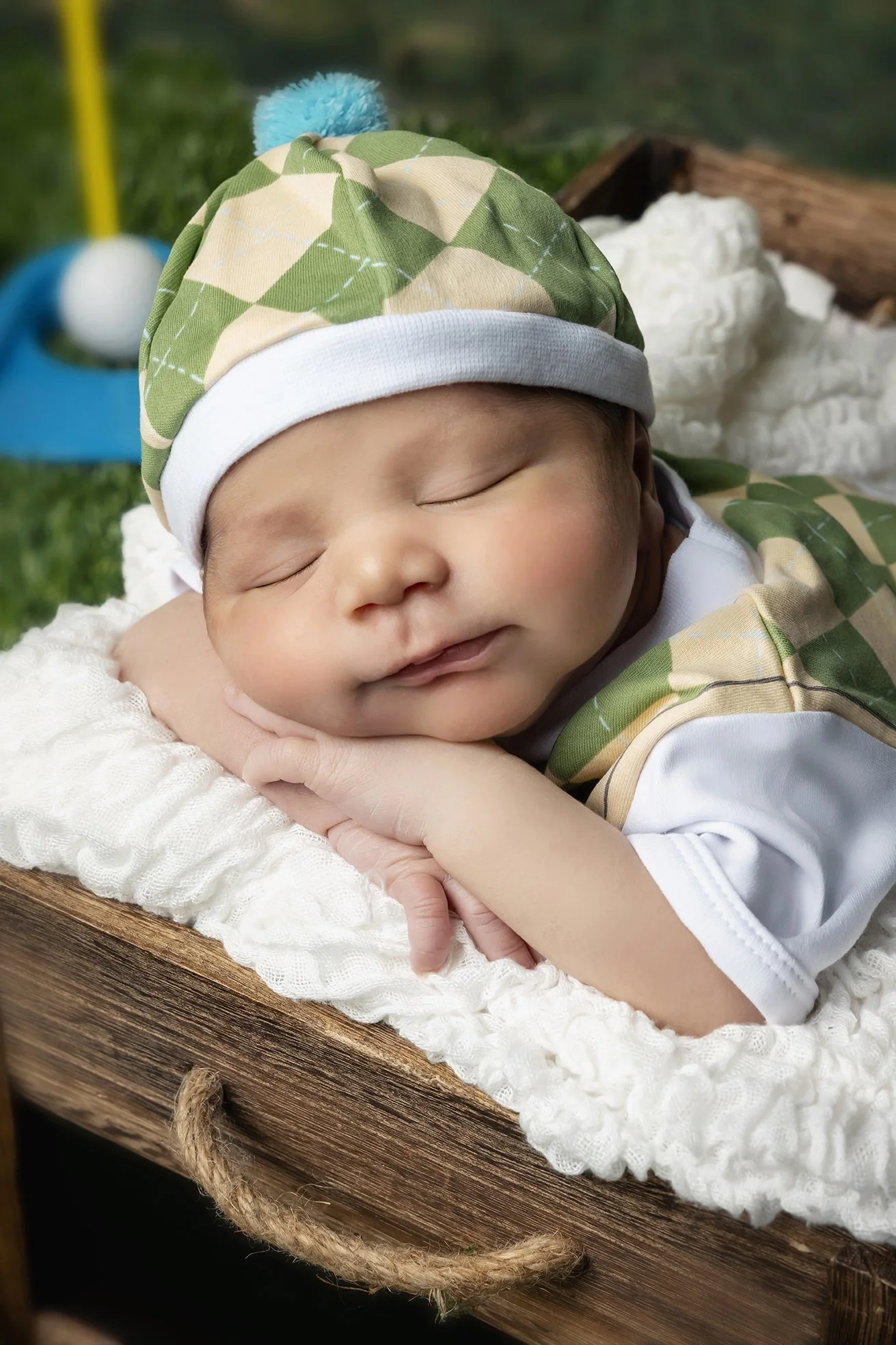 A sleeping baby dressed in camouflage clothes with a matching camouflage cap, lying on a soft white blanket in a rustic wooden cradle outdoors.