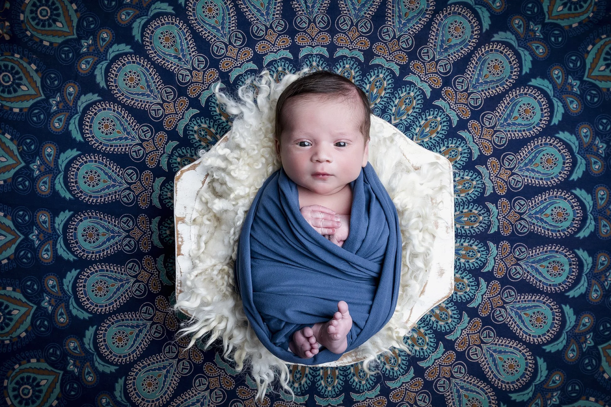 A baby wrapped in a blue blanket, lying on a white fluffy blanket in a white bowl on a patterned blue background.