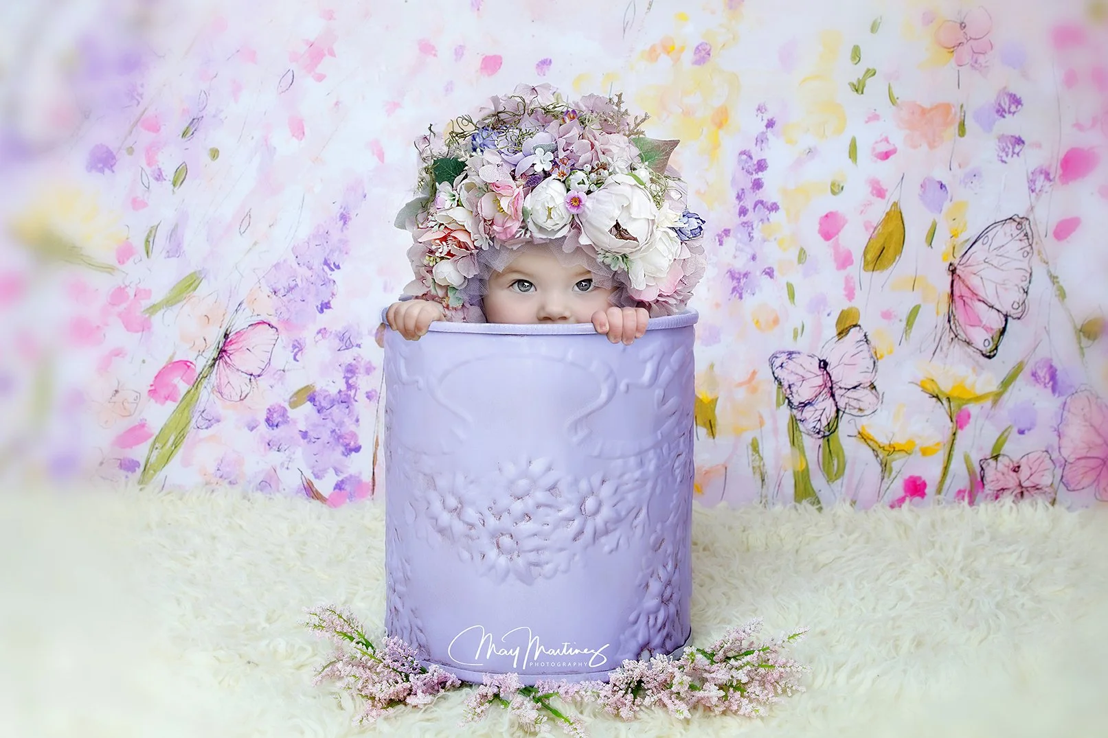 A child with blue eyes peeking out from behind a purple flower-covered container, wearing a floral crown, in a colorful, butterfly-themed setting.