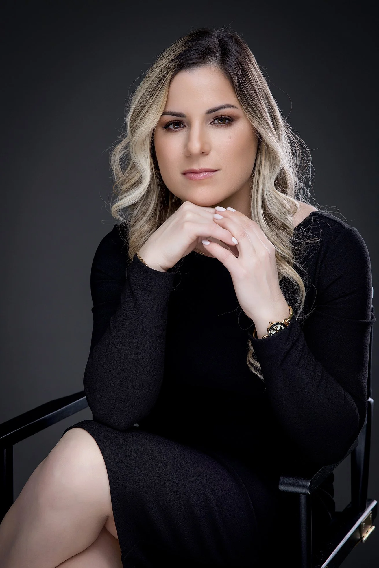 A woman with blonde wavy hair wearing a black long-sleeve dress sitting on a black chair against a dark background, resting her chin on her hands and looking at the camera.