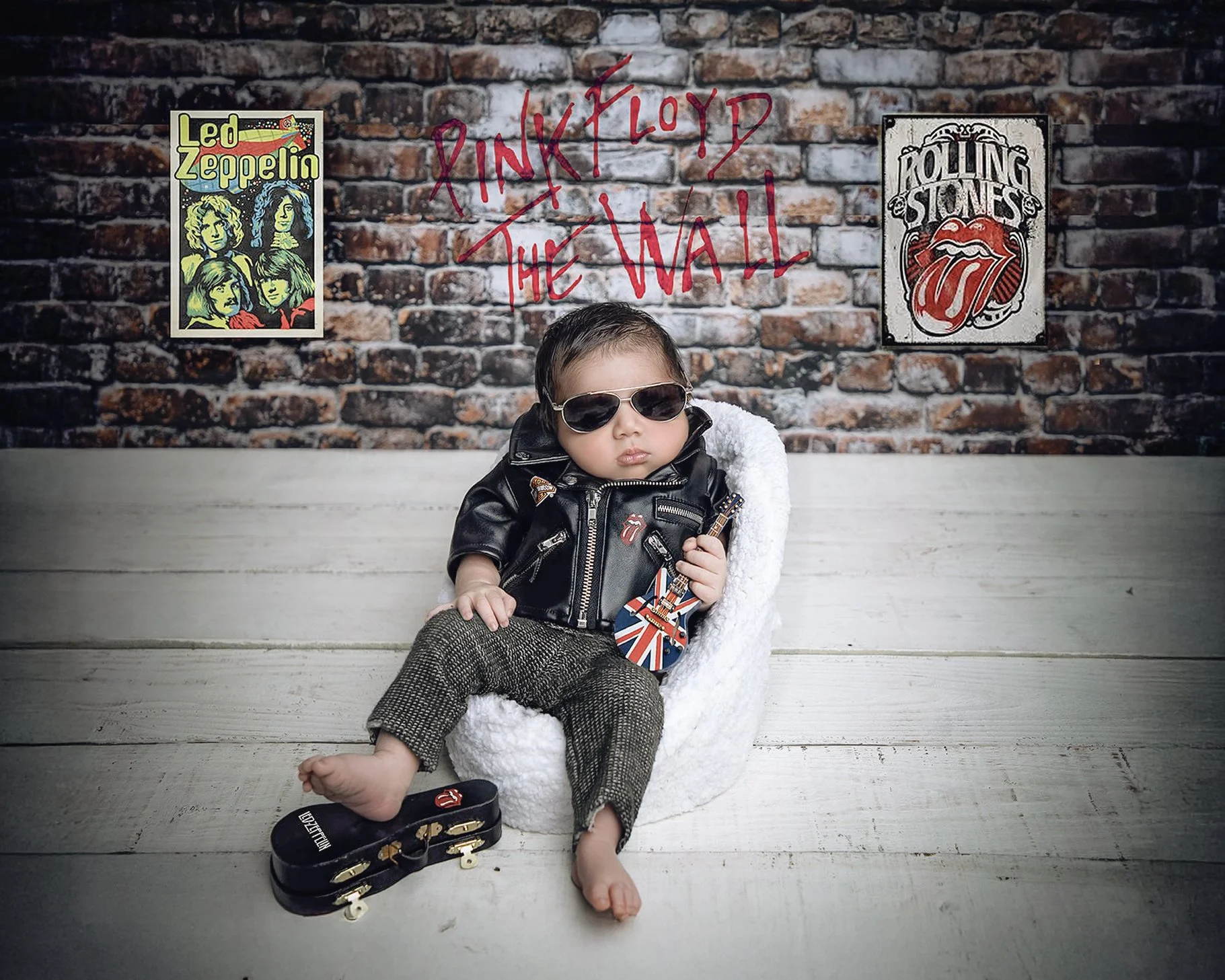 A young child with sunglasses, a black leather jacket, and patterned pants sitting on a white chair against a brick wall with 'Pink Floyd The Wall' written in red. Posters of Led Zeppelin and The Rolling Stones are on the wall next to the writing.