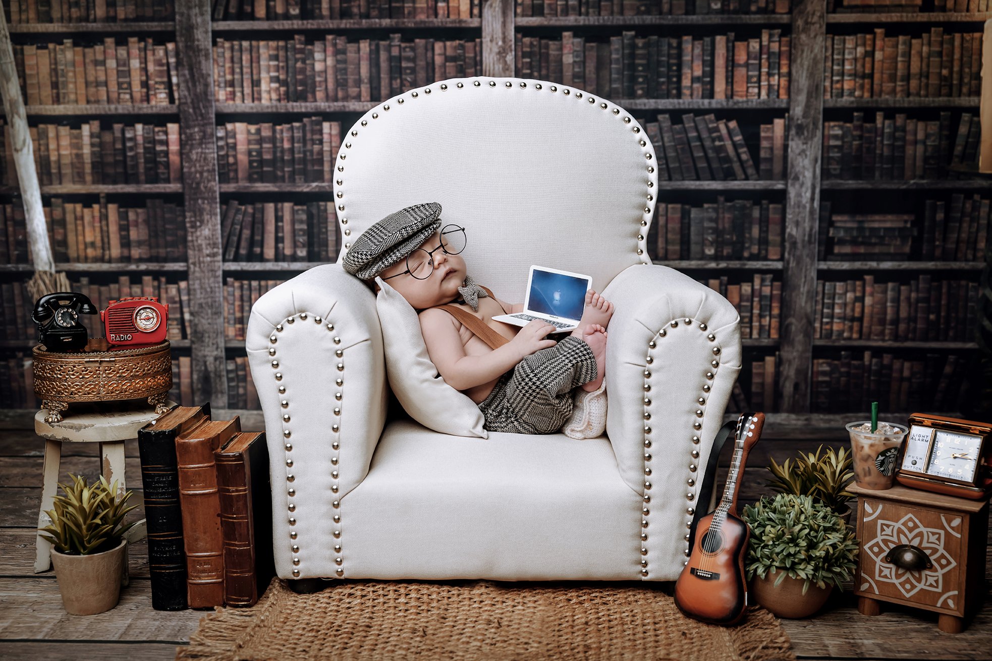 A small child dressed in vintage clothing and glasses, wearing a flat cap, sleeping in a large white armchair while holding a small laptop, in a cozy room with bookshelves, plants, a guitar, and vintage-style decor.
