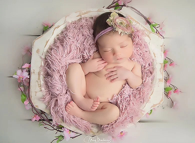 A sleeping baby girl with a floral headband, lying on a pink furry blanket inside a decorative bowl surrounded by pink flowers.