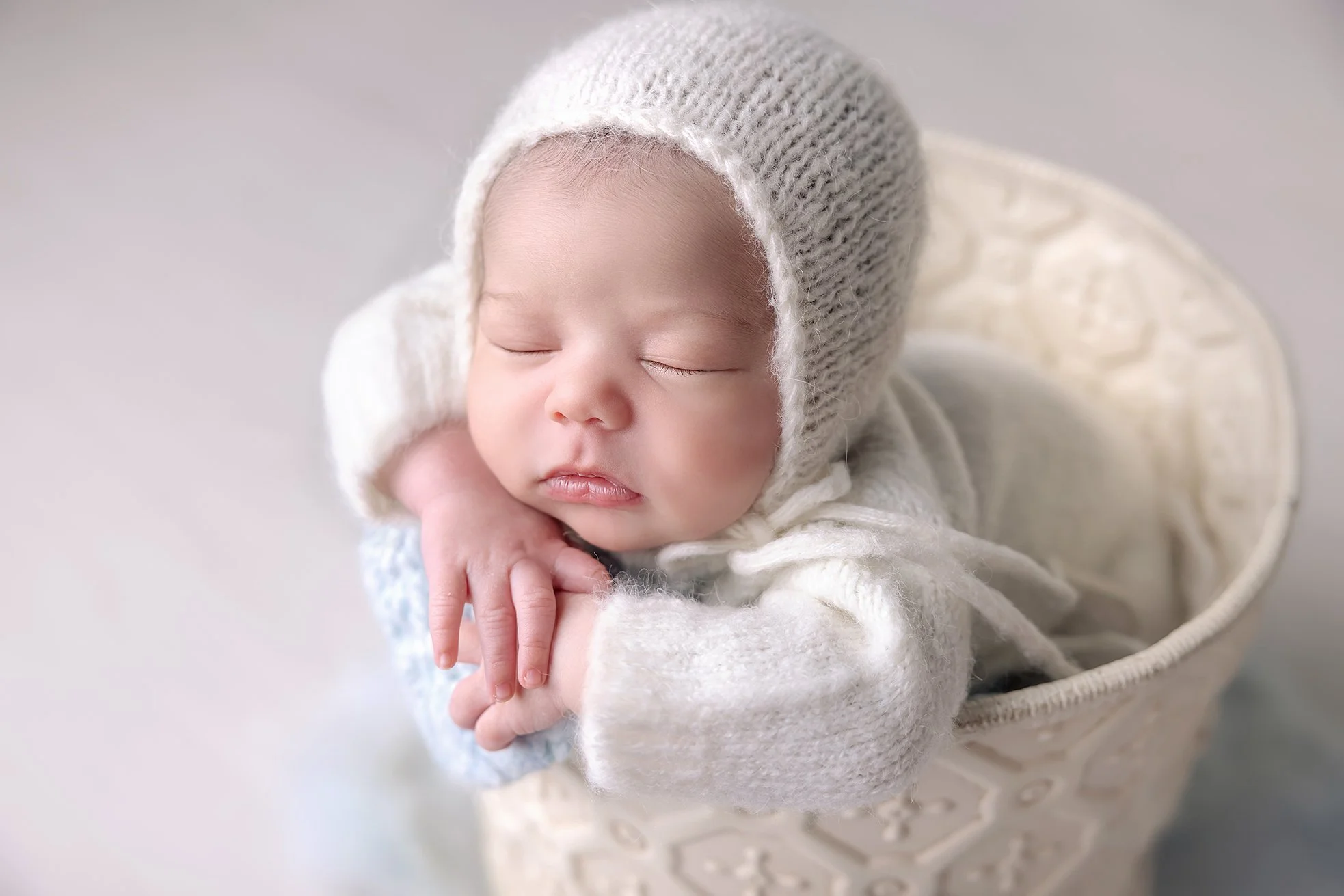 A sleeping baby with a knitted hat resting on their arm inside a cream-colored basket.