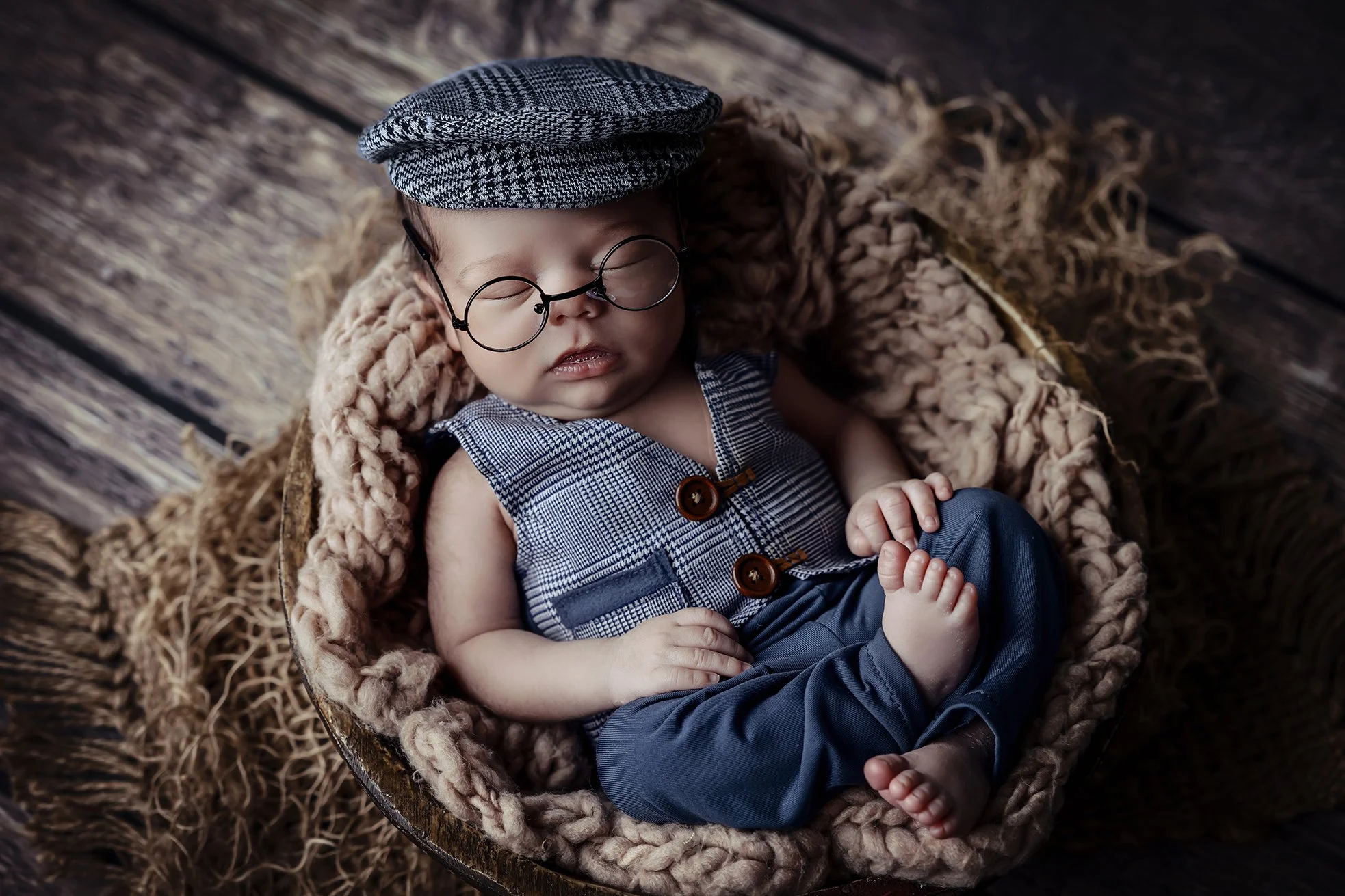 A baby dressed in vintage-style clothes, wearing round glasses and a checkered cap, sleeping in a rustic wooden basket lined with a soft, fluffy blanket, on a wooden floor.