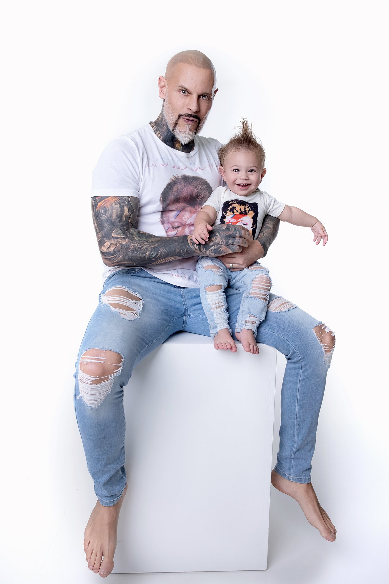 A man with tattoos wearing ripped jeans and a graphic t-shirt is sitting on a white block with a young girl on his lap. The girl has a quiff hairstyle, is smiling, and is wearing ripped jeans and a graphic t-shirt. They are against a plain white back