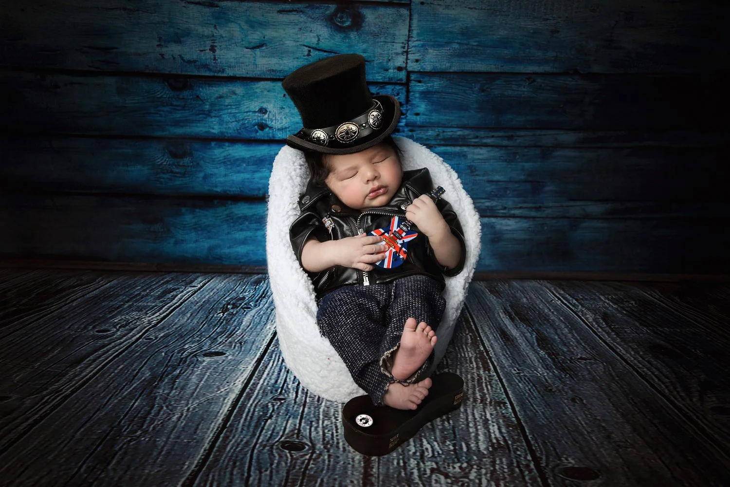 A sleeping baby wearing a leather jacket, a top hat with silver conchos, and plaid pants, holding a guitar pick with an Union Jack design, sitting in a white fuzzy chair against a blue wooden wall background.