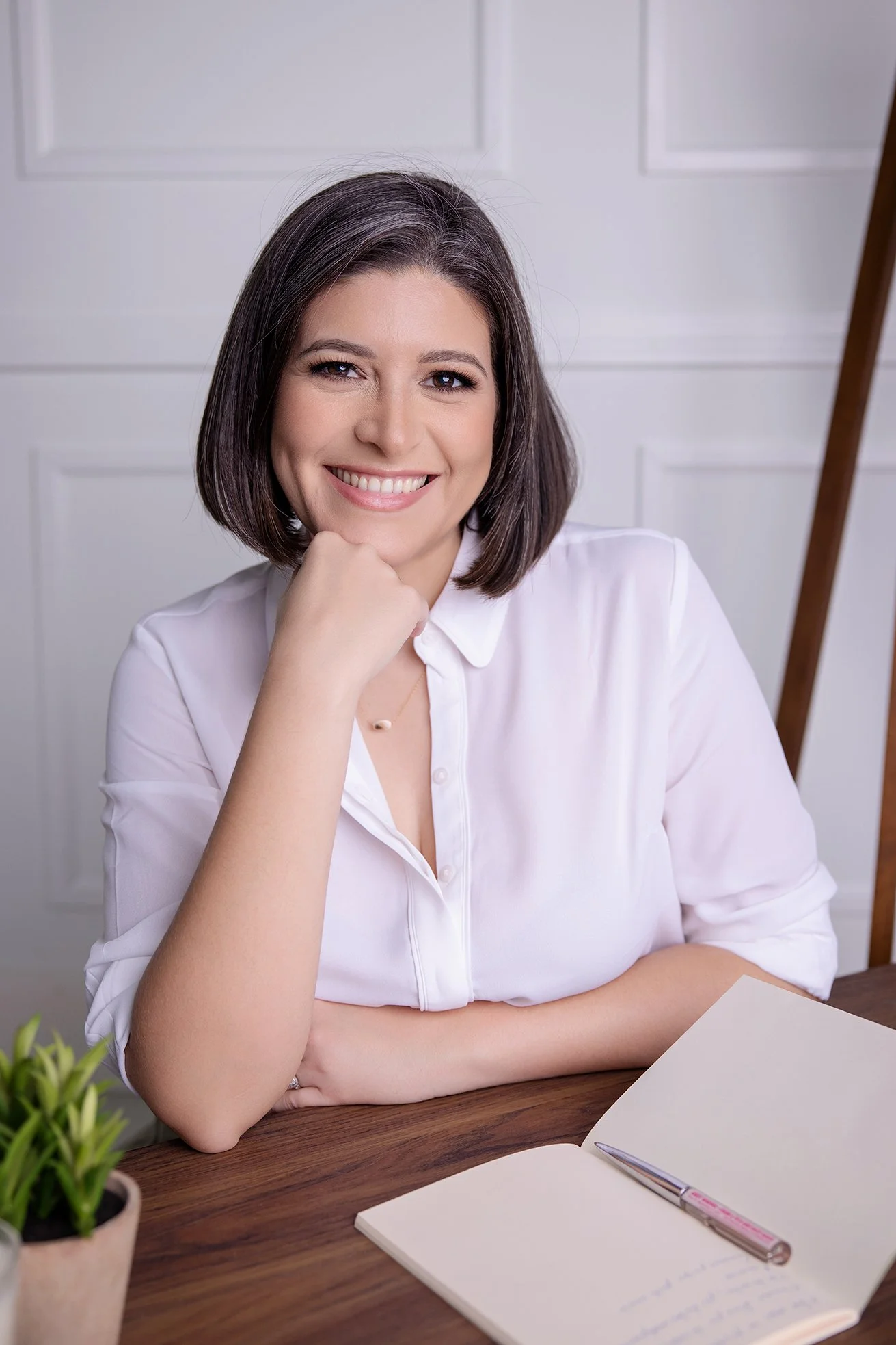 Smiling woman with dark brown hair sitting at a wooden table with an open notebook, pen, and potted plant in a white-walled room.