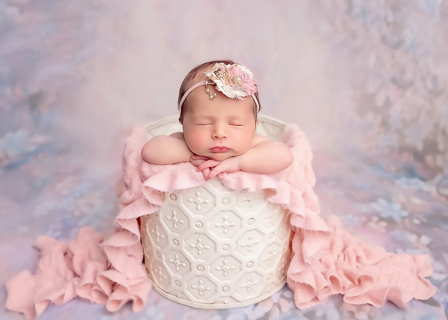A sleeping newborn baby with a pink headband and floral decoration, resting with arms folded on a ceramic container, draped in a pink blanket, against a soft pastel background.