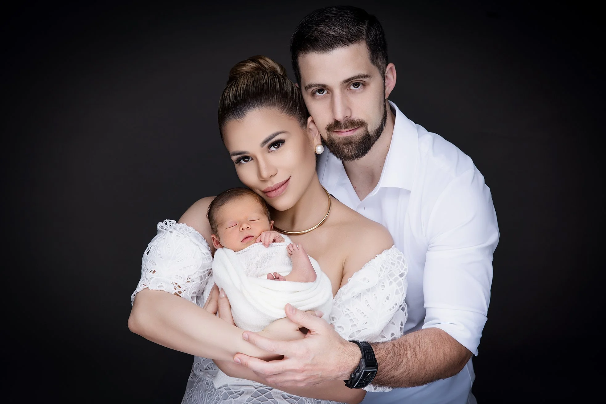 A family of three, a woman, a man, and a newborn baby, posing against a black background. The woman holds the sleeping baby wrapped in a white blanket, the man is standing behind her, and they are close together.