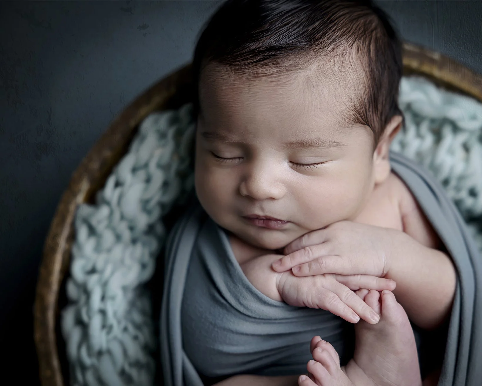 Close-up of a sleeping baby with closed eyes, resting in a basket with hands gently resting on the face.