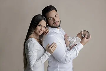 Family of three with a woman, man, and baby against a neutral background.