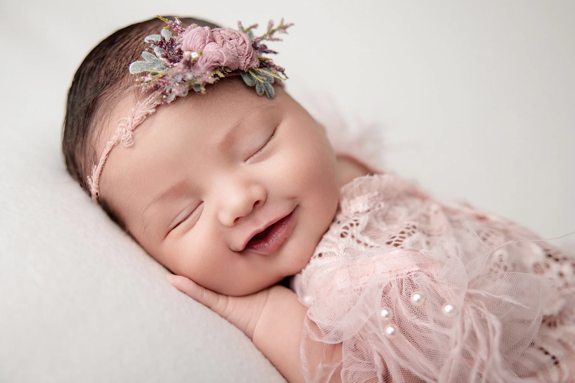 Smiling baby girl with a pink floral headband and lace dress, resting on a soft surface.