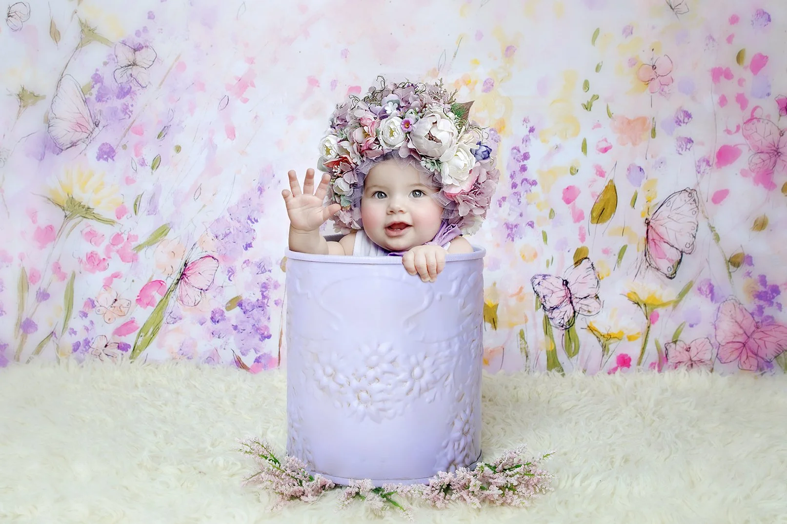 A baby with blue eyes and rosy cheeks, wearing a large floral headpiece, sitting inside a white decorative container, waving, against a colorful floral and butterfly decorated background.