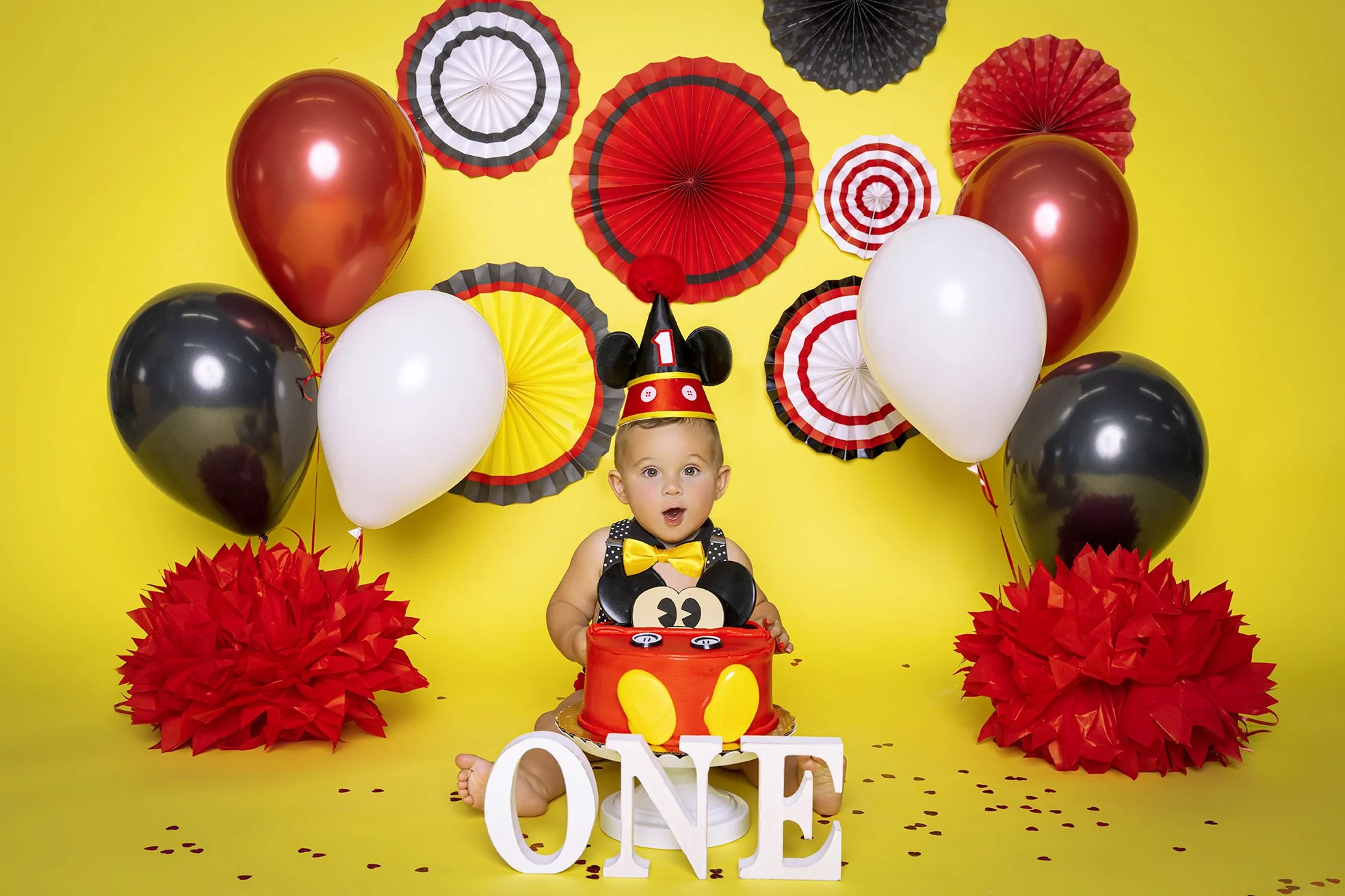 A baby celebrating their first birthday with a Mickey Mouse themed cake, party hat, and decorations including balloons and paper fans on a yellow background.