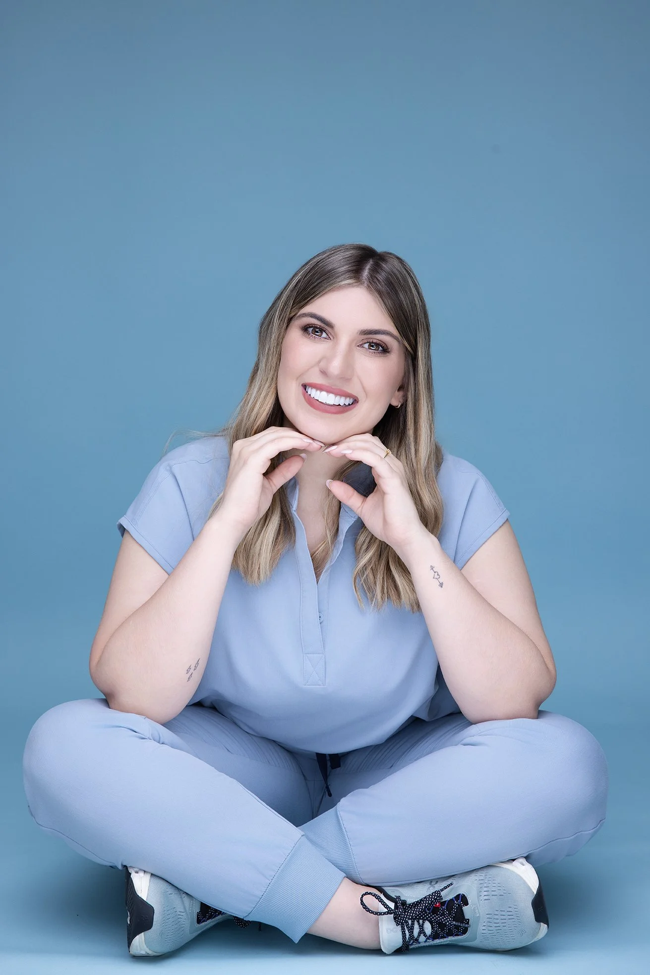 Young woman sitting cross-legged on the floor, smiling happily, with her hands framing her face against a solid light blue background. She is wearing a light blue casual outfit and sneakers.