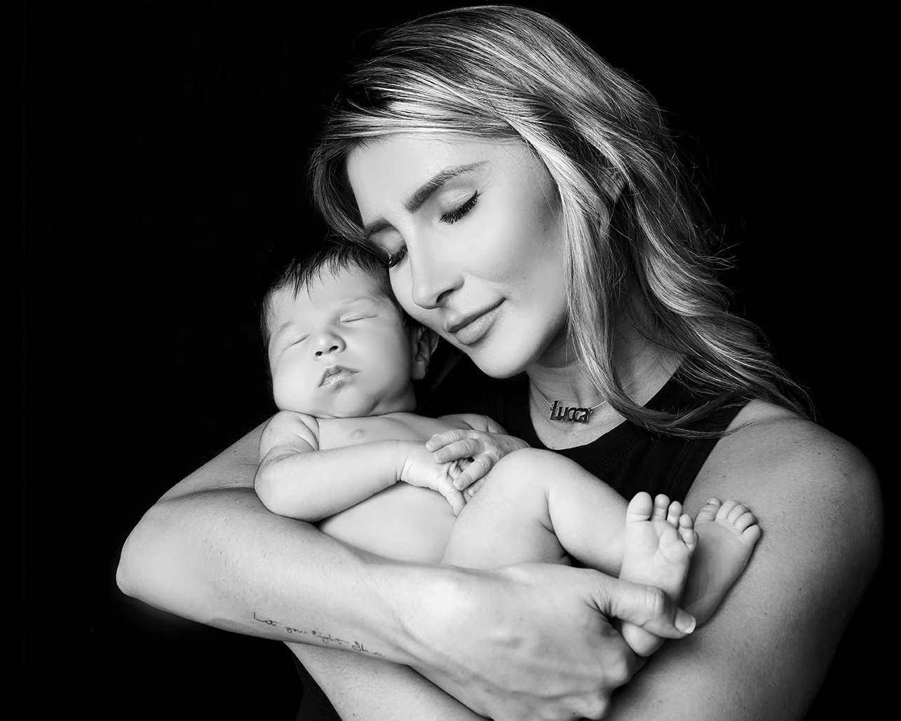 A woman is gently holding a sleeping baby close to her chest. They are both peacefully resting, with the woman smiling softly and her eyes closed. The image is in black and white.