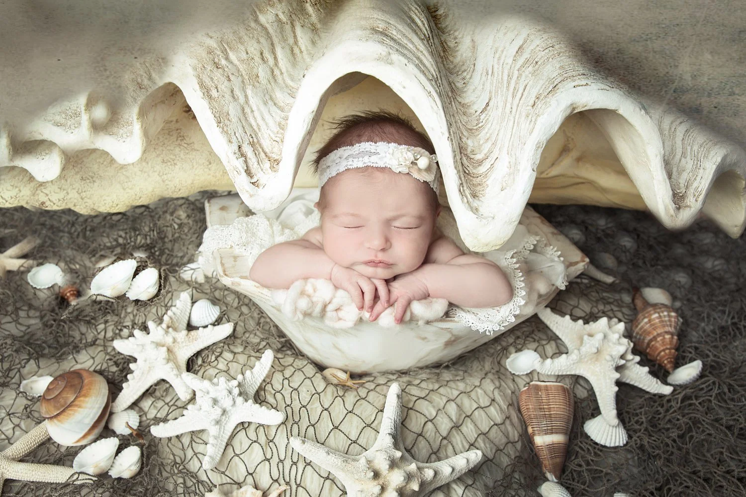 A sleeping baby with a headband, lying on a bed of shells and starfish under a large seashell.