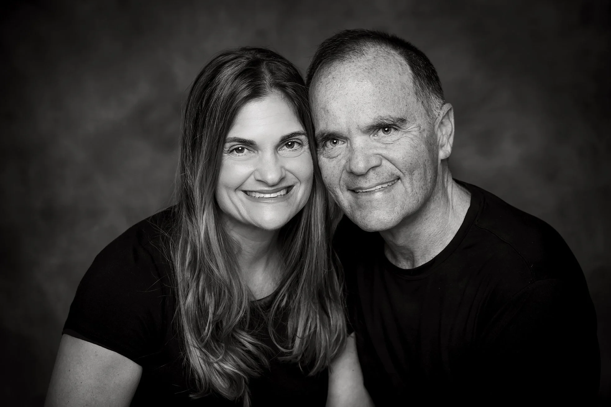A black and white portrait of a smiling woman with long wavy hair and a man with short hair, both wearing black shirts, close together against a dark textured background.