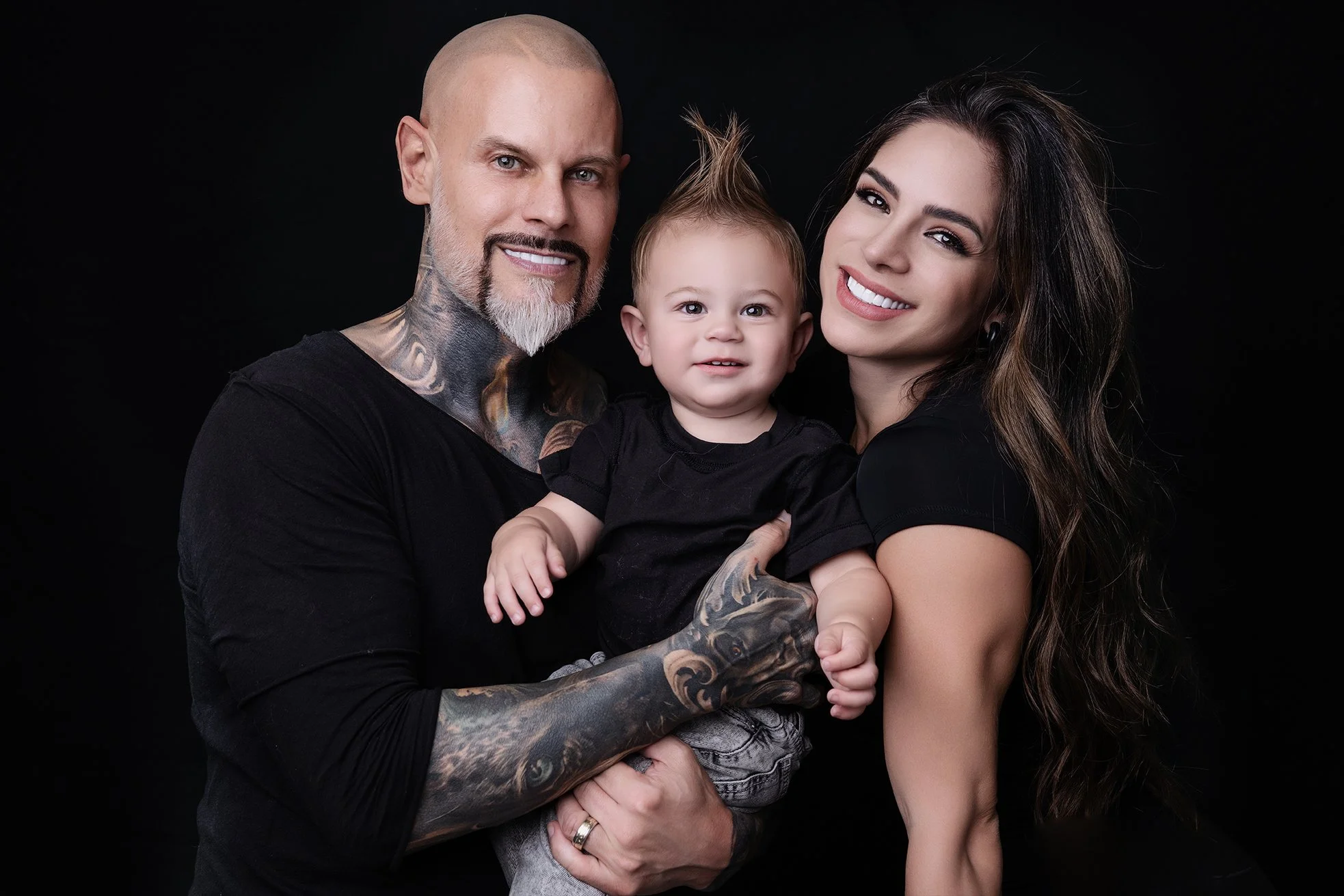 A smiling family portrait featuring a tattooed man with a bald head, a woman with long wavy hair, and a young child with a mohawk, all wearing black shirts and posing together against a black background.