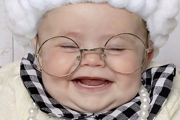 A smiling baby wearing large round glasses, a fluffy white hat, and a checkered outfit with pearls.