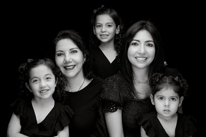 A black and white photo of two women and three young girls, all smiling and dressed in dark clothing, posing closely together against a dark background.