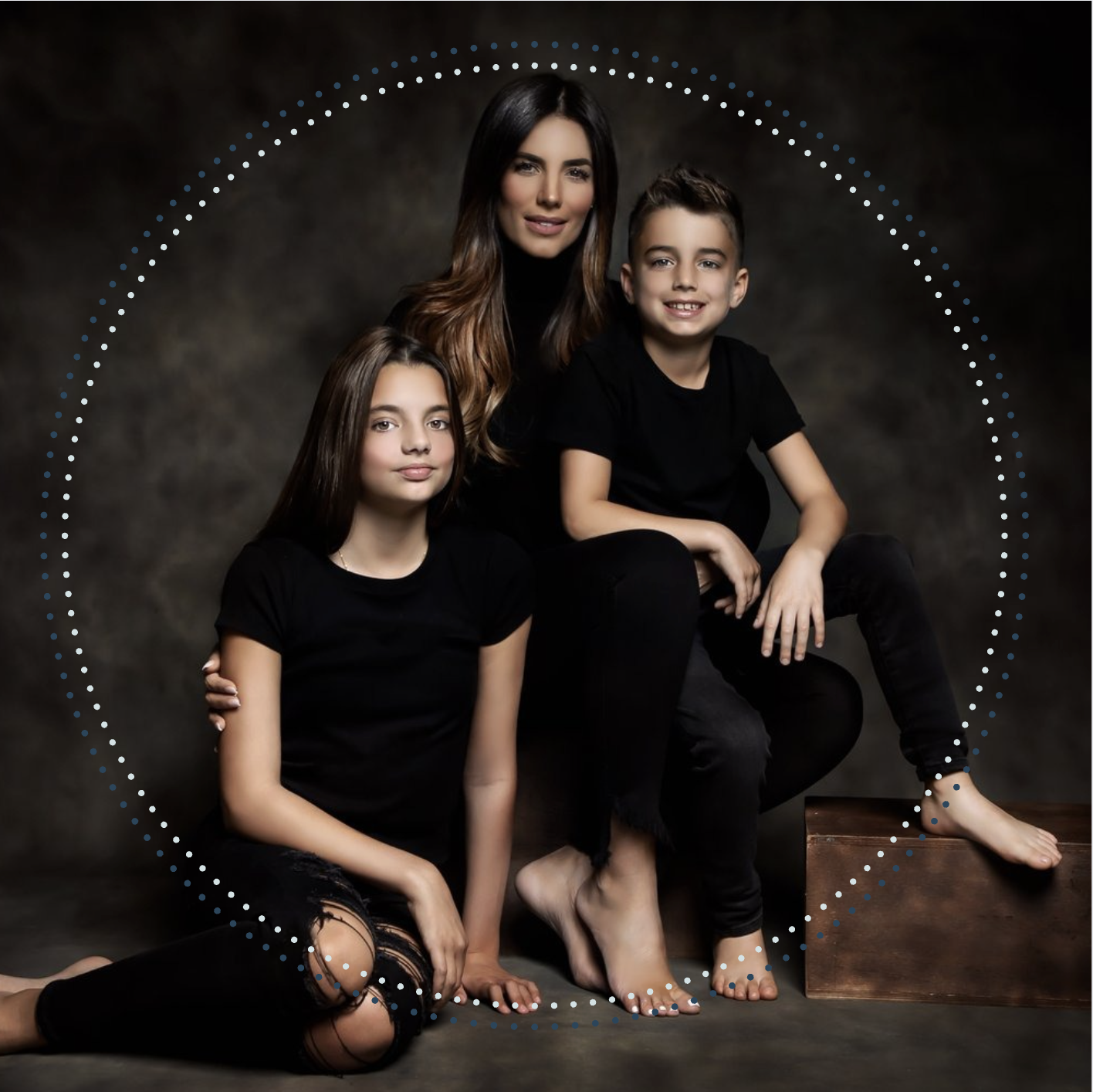 A woman and two children, a girl and a boy, pose together against a dark backdrop. They are dressed in black, with the woman sitting on a wooden box and the children sitting or kneeling nearby. The girl has long brown hair, and the boy has short hair. The woman has long, wavy brown hair.