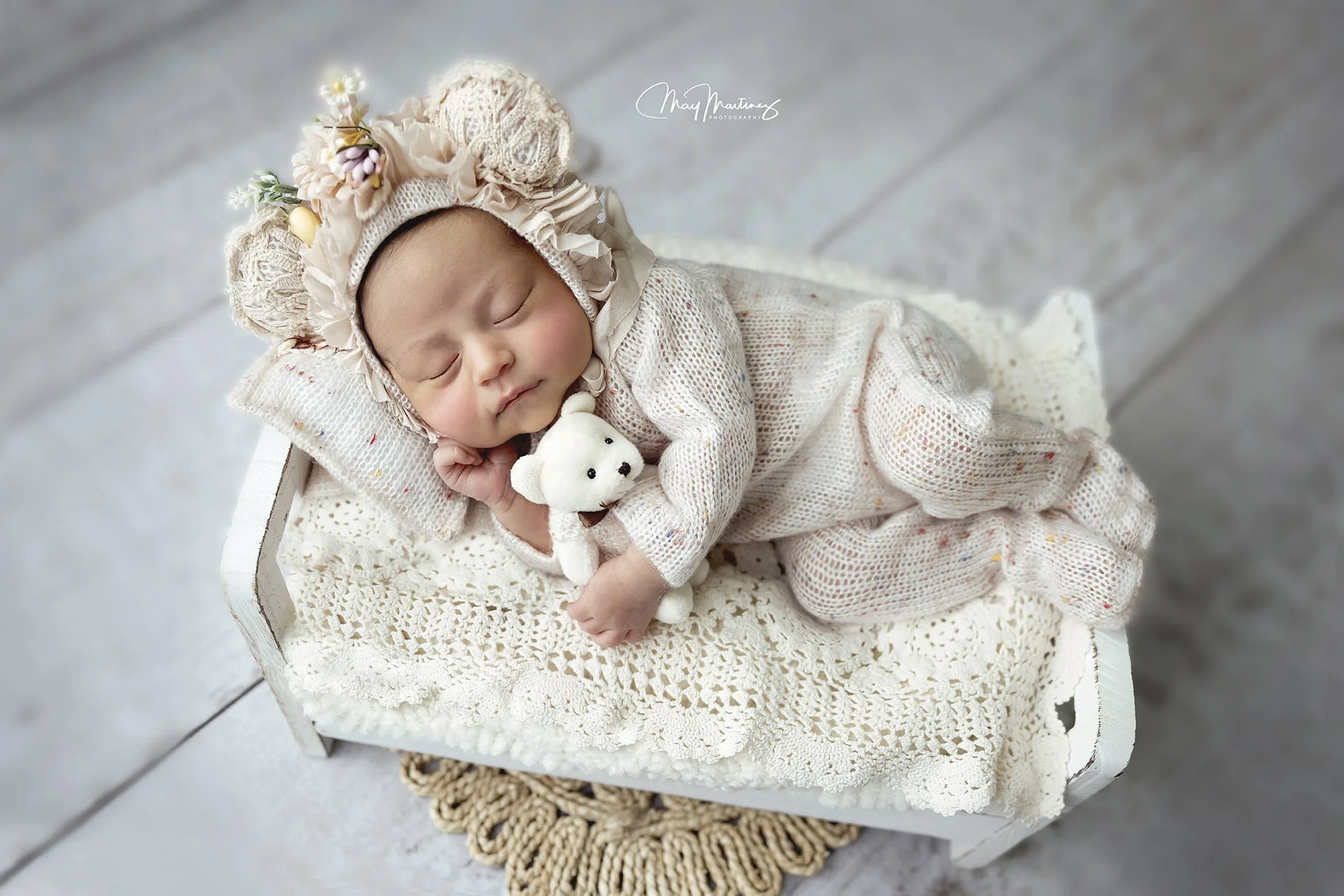 A sleeping baby lying on a crochet blanket in a vintage white wooden crib, wearing a knit hat with flowers and bear ears, holding a small white teddy bear, on a light gray wooden floor.