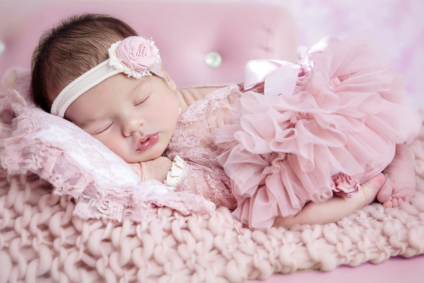 A sleeping baby girl lies on a pink knitted blanket, dressed in pink lace and tulle frilly dress with a matching headband featuring a pink flower.