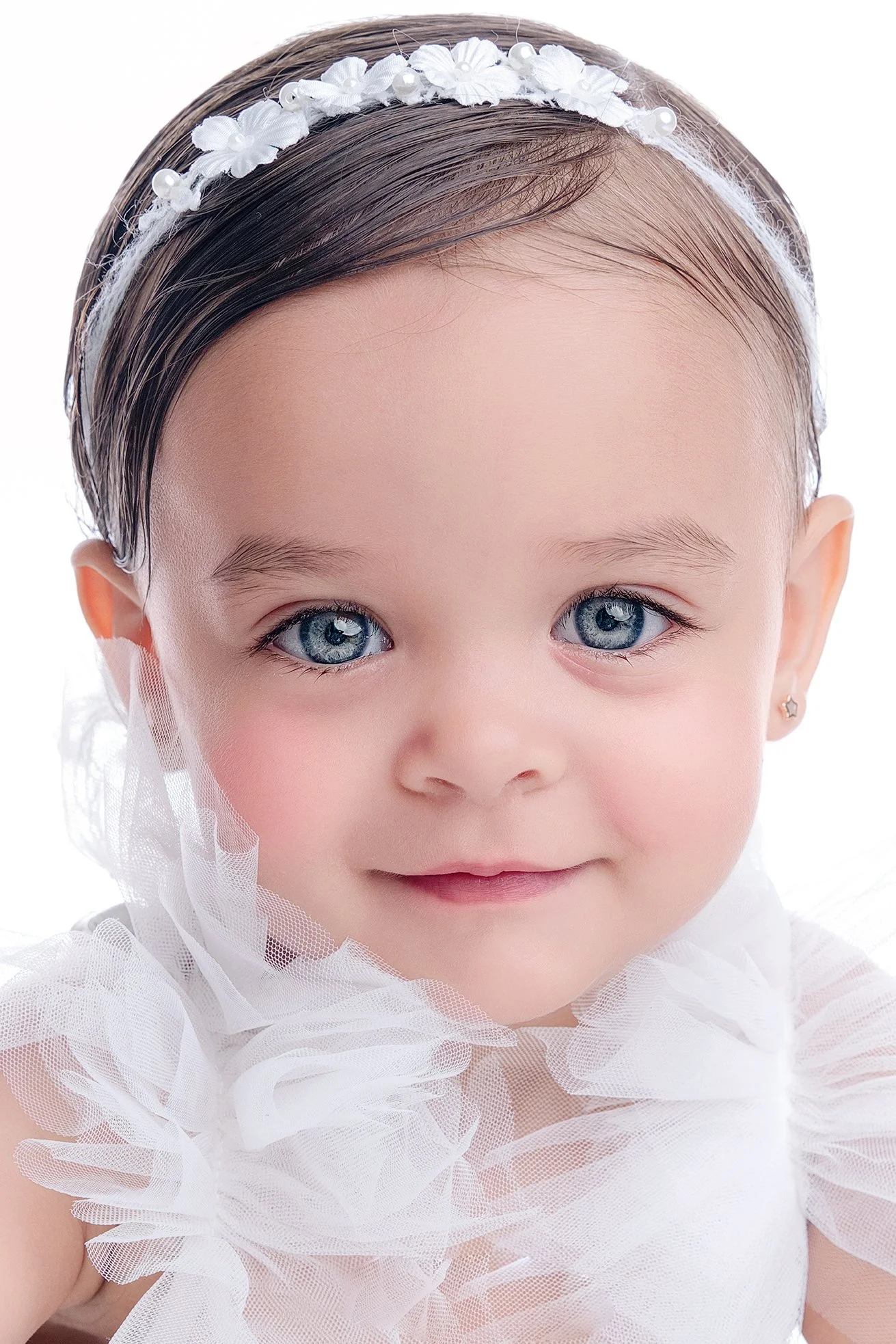 Close-up of a young girl with bright blue eyes, wearing a white headband with flowers and pearls, and a white ruffled dress.