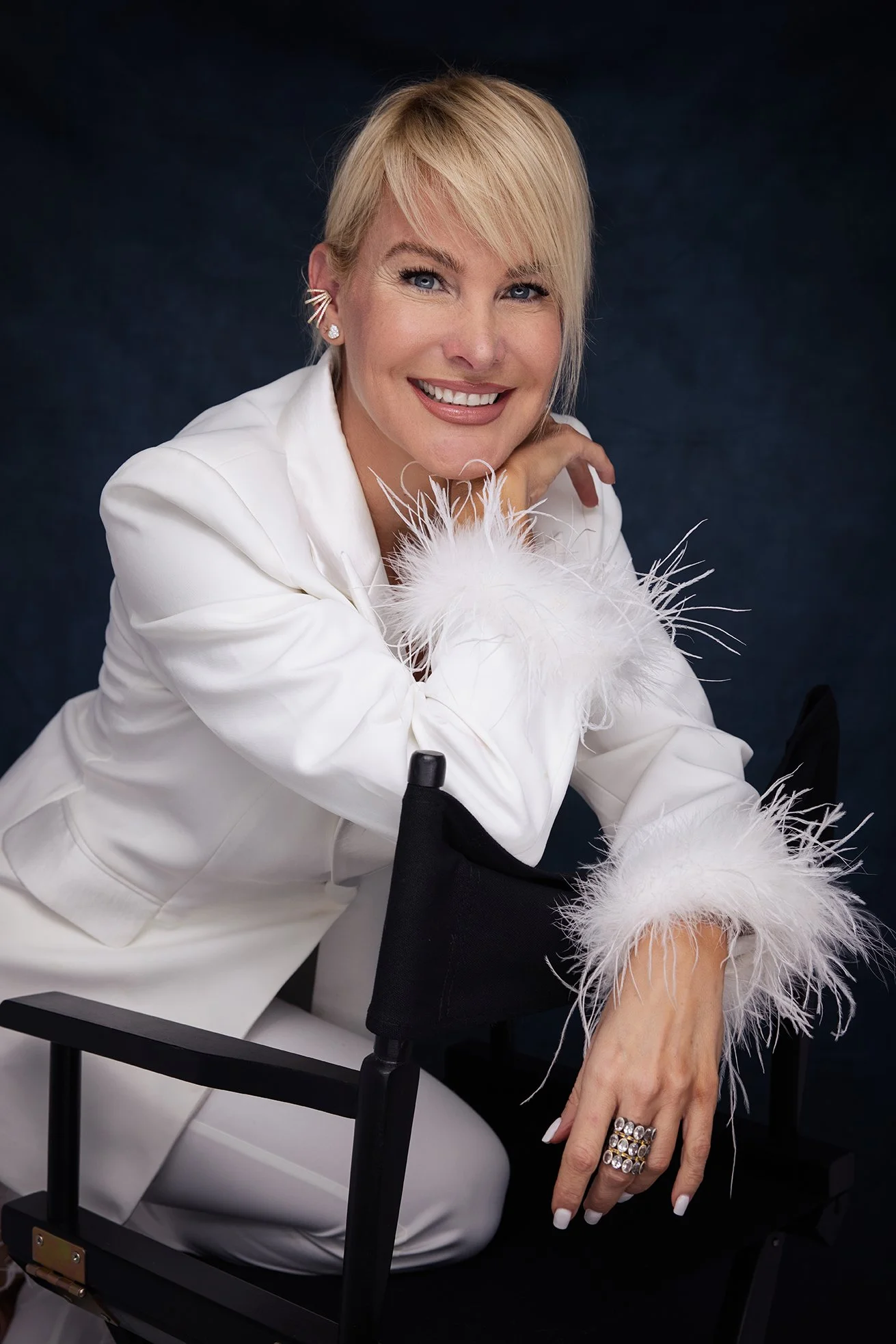 A woman with blonde hair and blue eyes, smiling while sitting on a director's chair, wearing a white suit with feathered cuffs and jewelry, against a dark background.