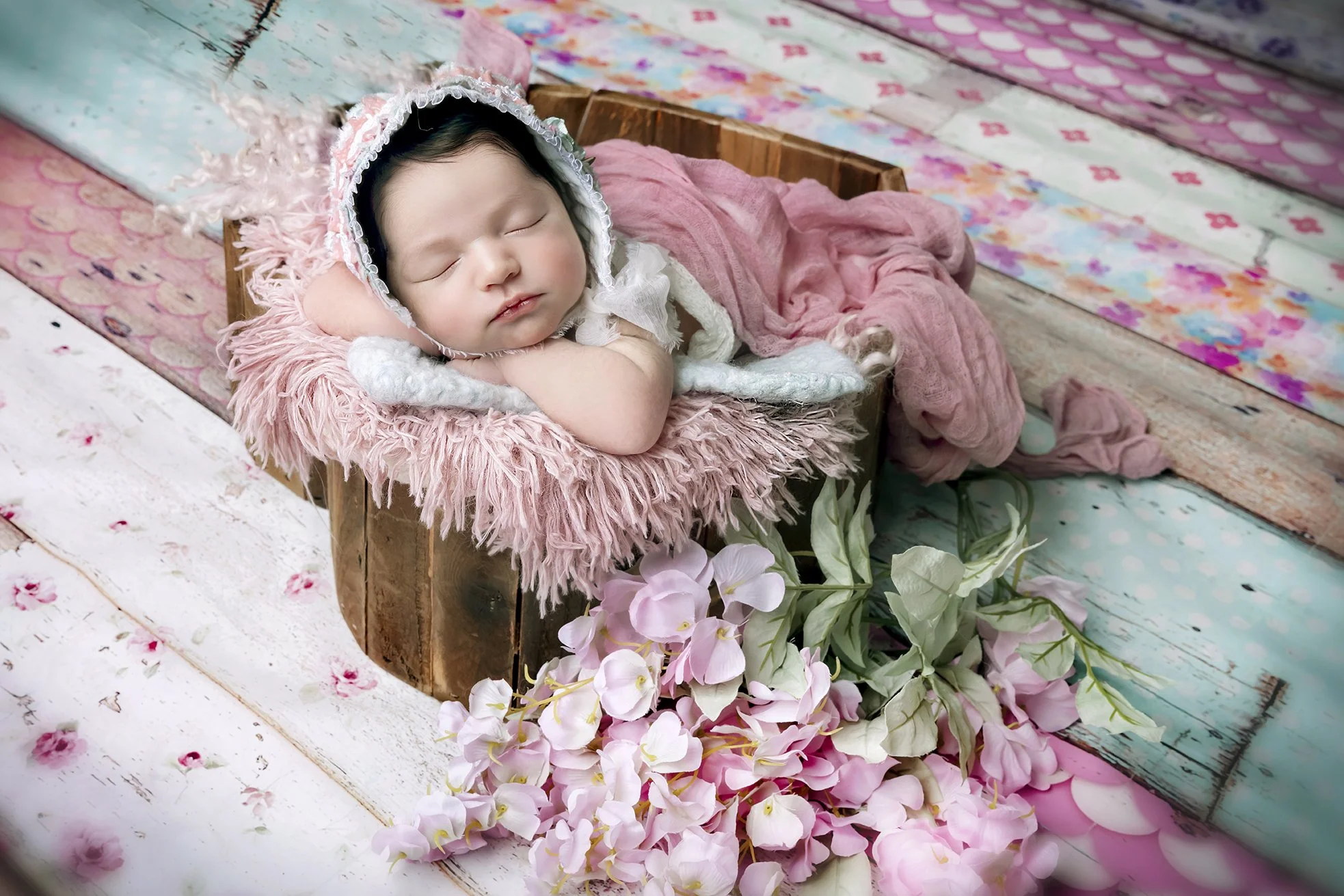 A sleeping baby girl wearing a bonnet, resting on a pink fuzzy blanket in a small wooden box surrounded by pink flowers and pastel-colored wooden planks.