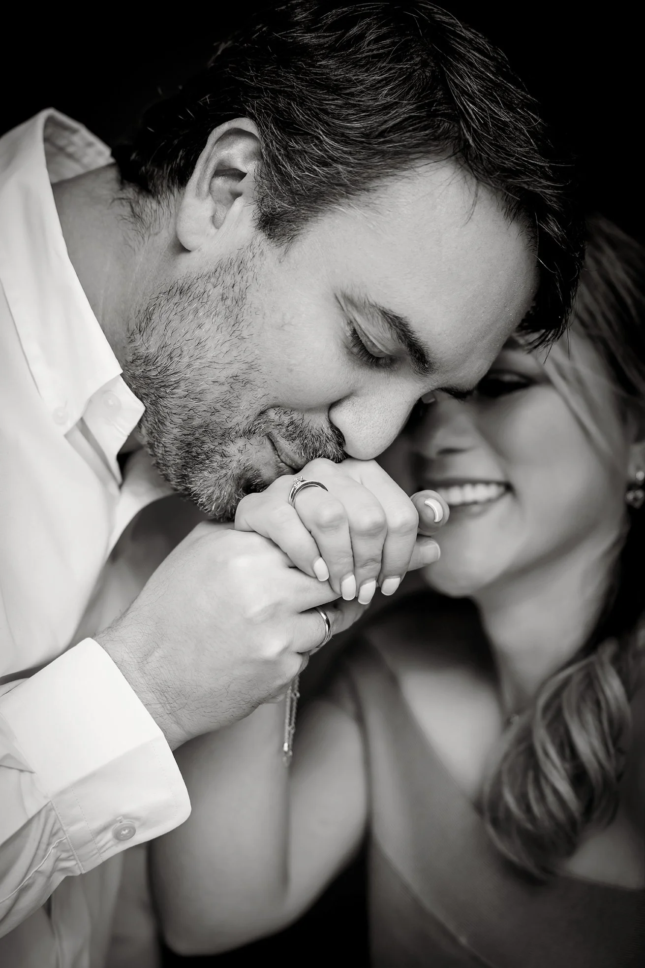 A black-and-white close-up photograph of a man and woman sharing an intimate moment, with the man kissing the woman's hand and both smiling.