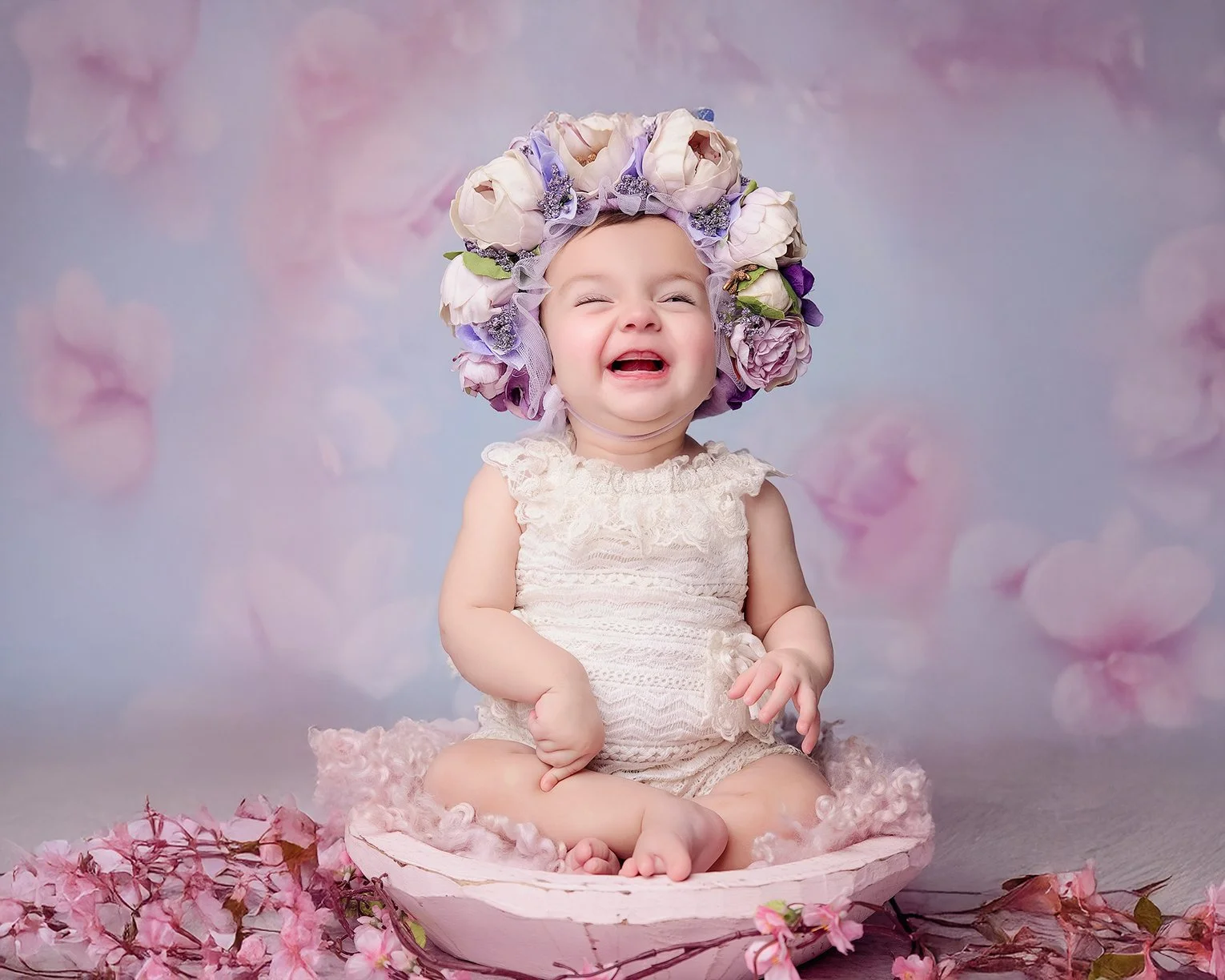A baby girl sitting in a pink bowl surrounded by pink flowers, wearing a cream-colored lace dress and a large floral headpiece, smiling and crying.