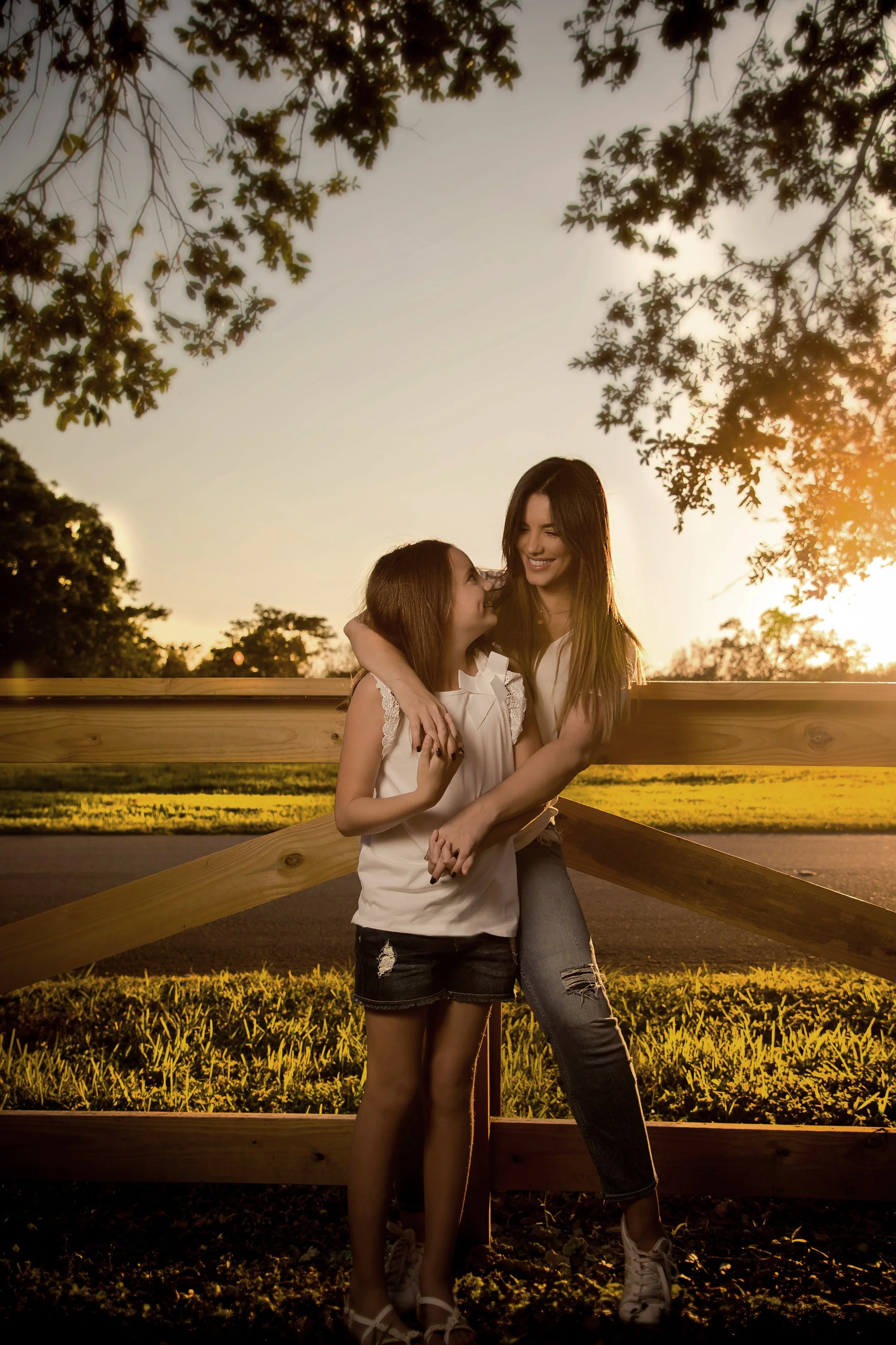 A woman and a girl standing outdoors near a wooden fence during sunset, smiling and hugging each other.