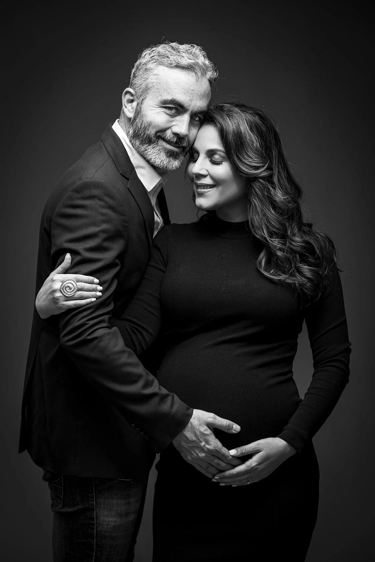 Black and white portrait of a couple, with the man embracing the pregnant woman, both smiling softly and looking happy.