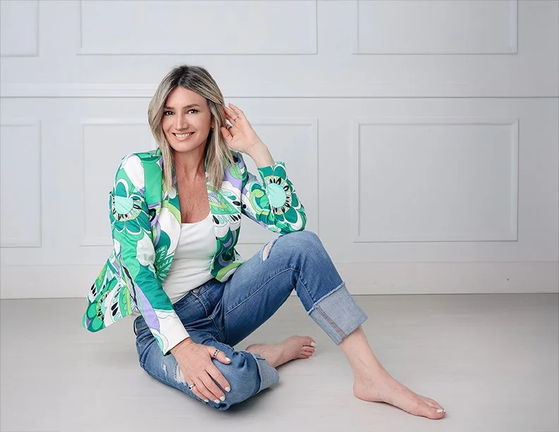 woman sitting on the floor in front of white paneled wall, smiling, wearing a vibrant patterned blazer, white top, and rolled-up jeans