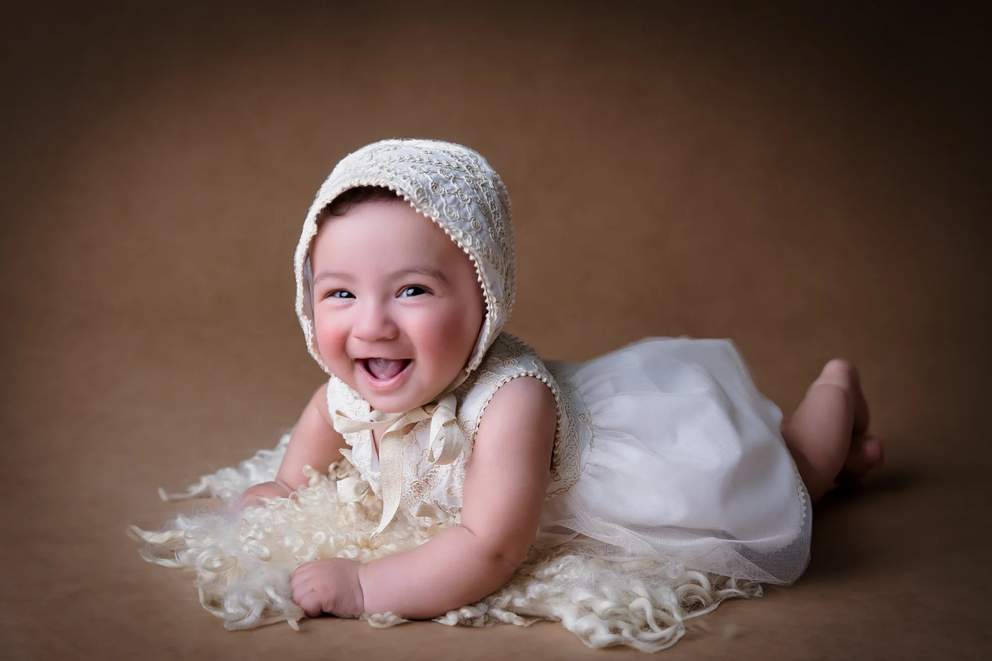 Smiling baby girl lying on a furry blanket, wearing a vintage lace bonnet and a white dress.