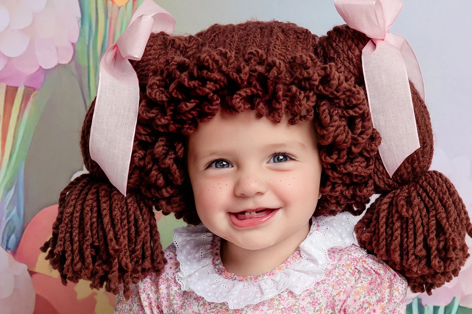 A young girl with curly brown hair, blue eyes, and freckles, smiling and wearing a pink floral dress with a white lace collar. She has large, pink fabric bows on either side of her head.