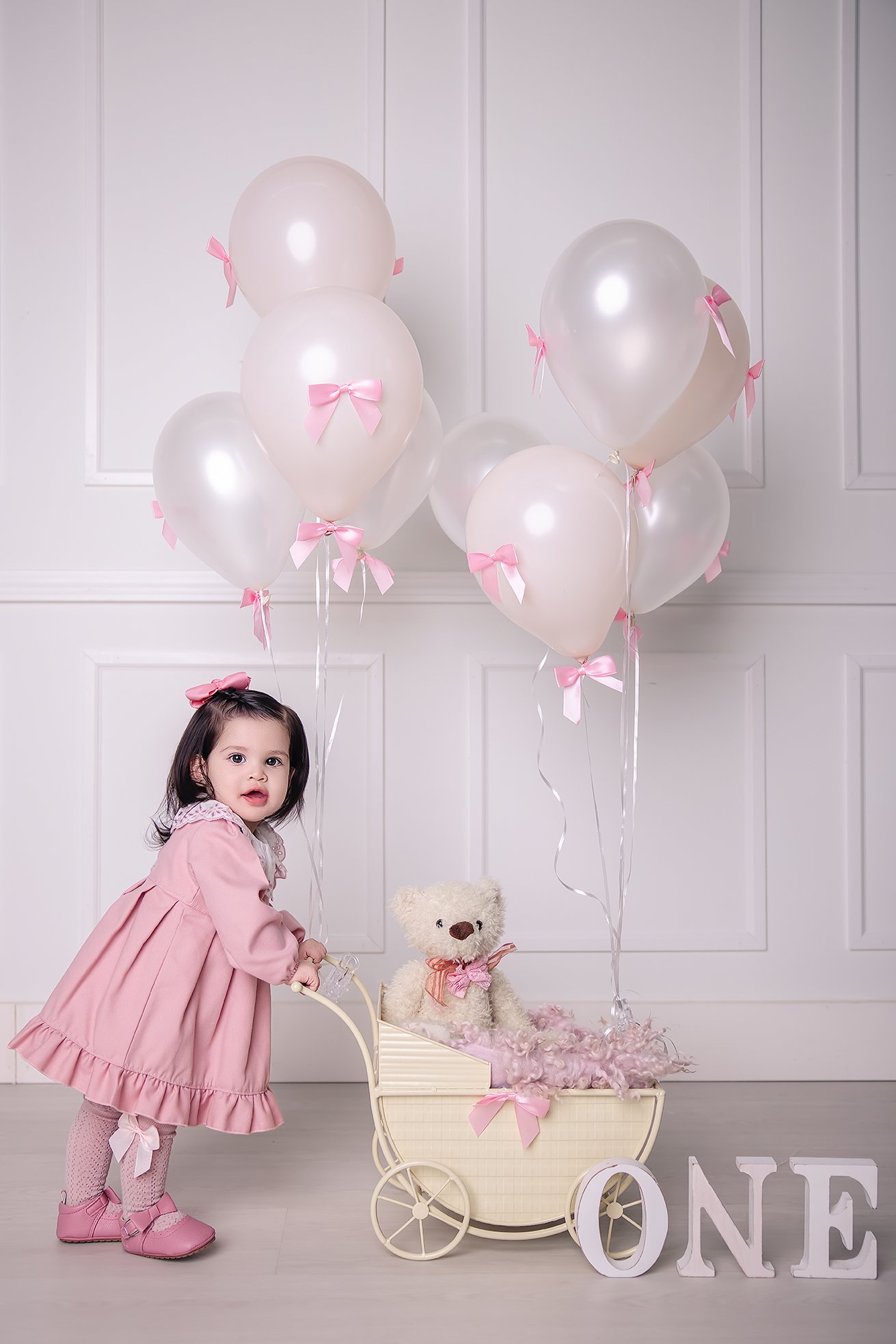A young girl in a pink dress and matching shoes stands next to a vintage-style toy stroller decorated with pink ribbons, holding a teddy bear, with pink balloons and large white letters spelling "ONE" in front, celebrating her first birthday.