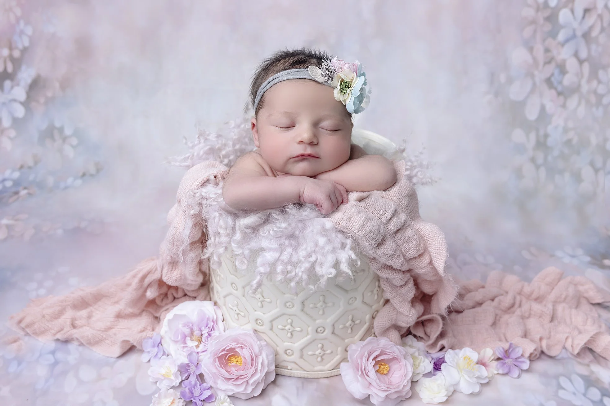 A sleeping baby with a floral headband, resting on a plush blanket in a decorative white vase, surrounded by pink and purple flowers.