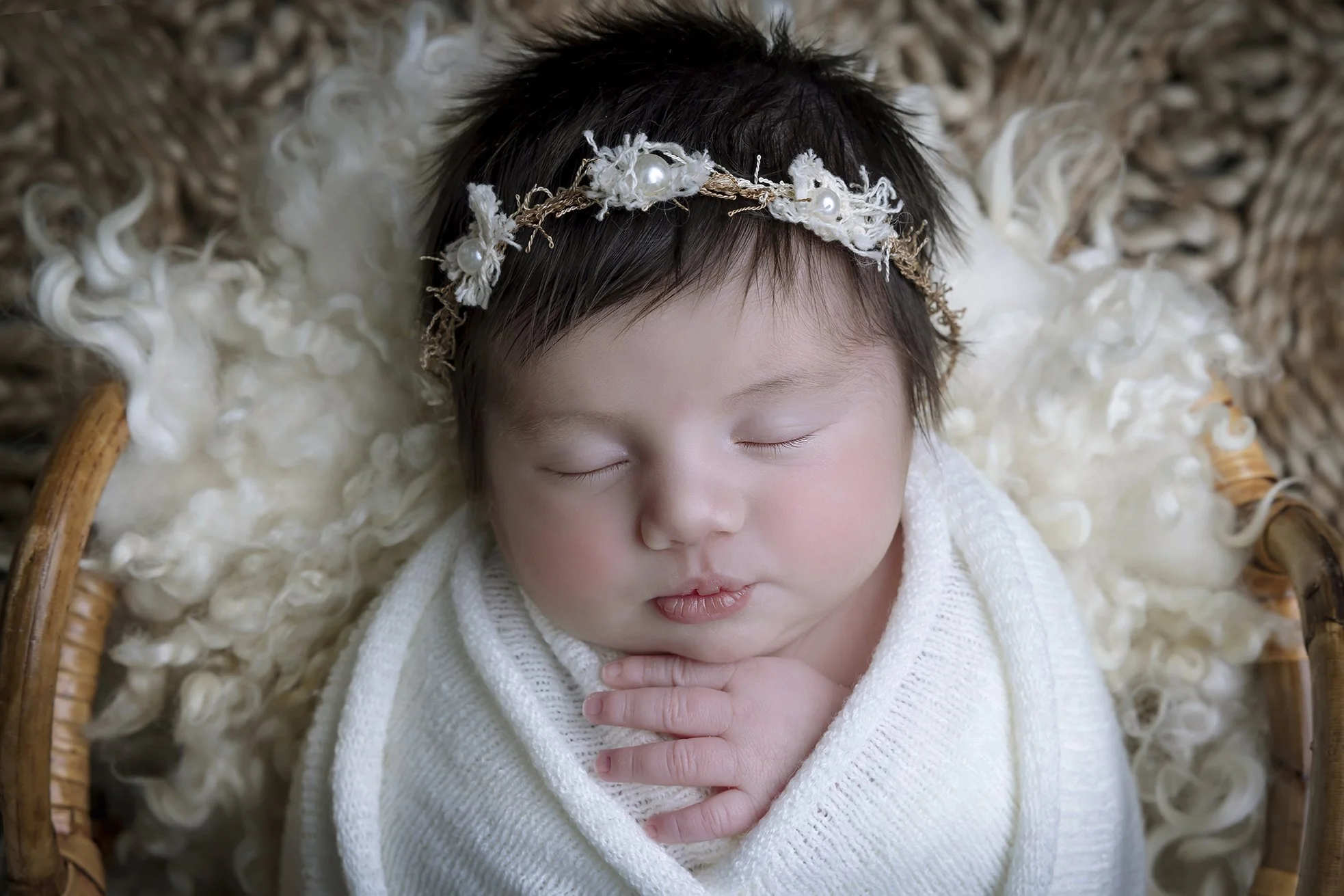 A sleeping baby with a floral headband, wrapped in a white blanket, lying in a wicker basket on a fluffy cream-colored blanket.