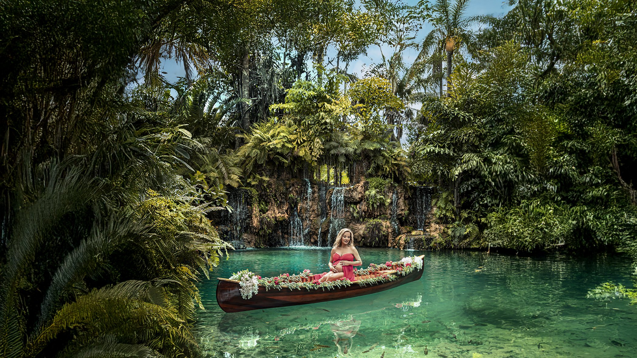 A woman in a red dress sitting in a floral decorated boat on a tropical turquoise river with waterfall and lush green trees in the background.