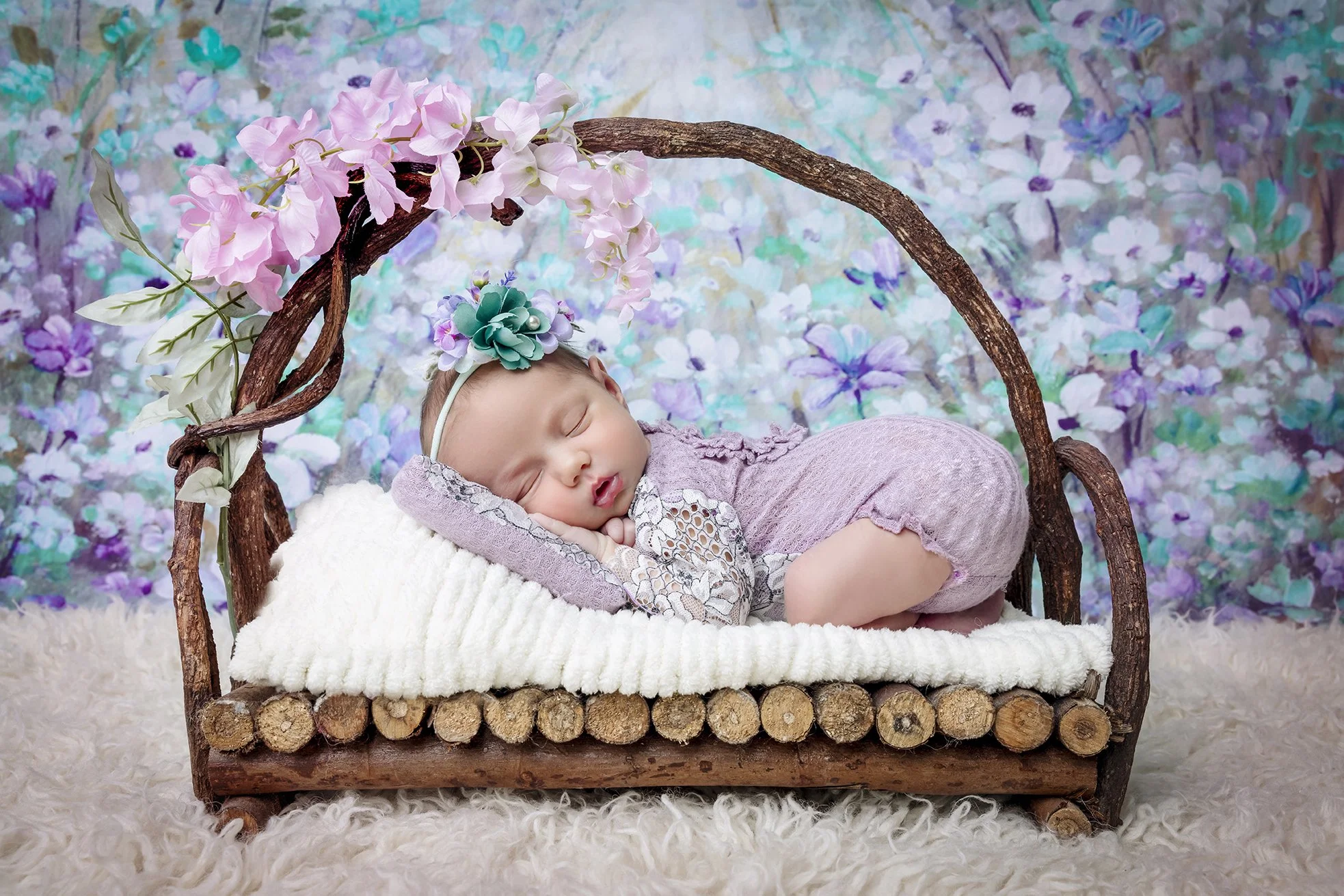 A sleeping baby girl with a floral headband lying on a small bed with a log frame and pink flowers, against a colorful, painted floral background.