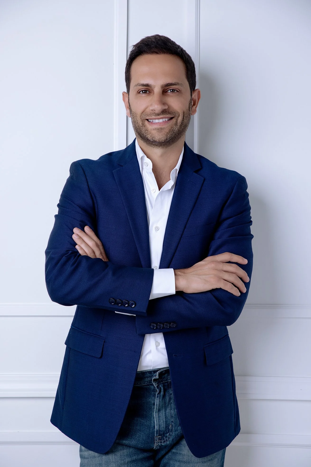 A man in a blue blazer and white shirt with arms crossed, standing against a white background, smiling.