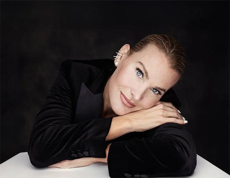 Portrait of a woman with short hair and blue eyes resting her head on her arms, wearing a black blazer against a dark background.