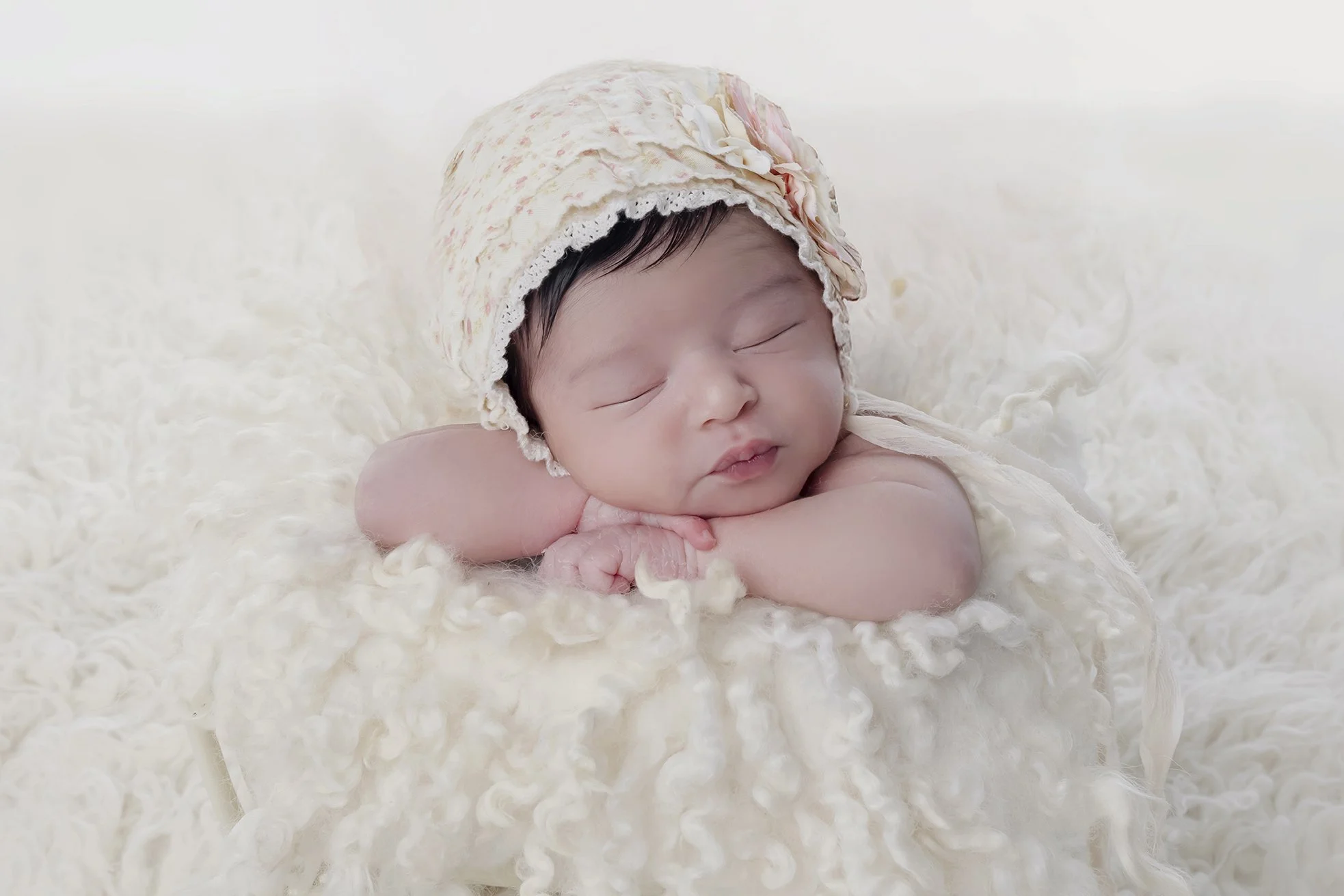 A sleeping baby with a floral bonnet, resting on a soft, cream-colored blanket.