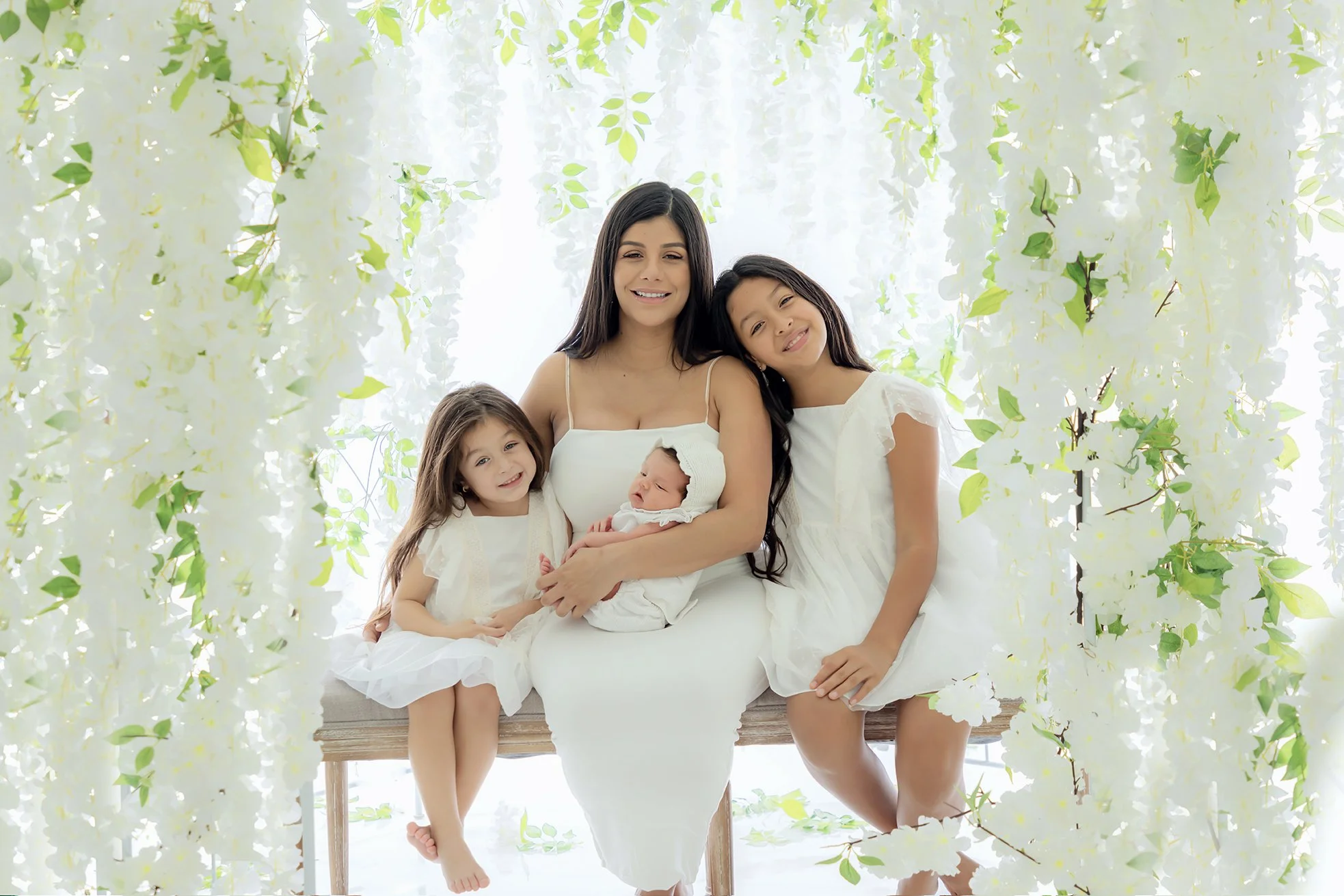A woman with three young girls, including a newborn, sitting on a bench surrounded by white flowers and green leaves.