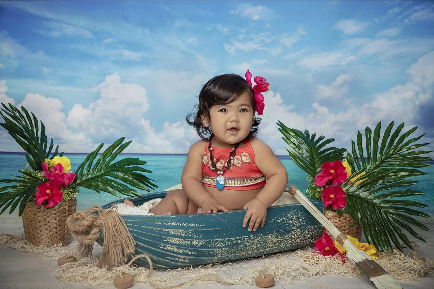 A young girl dressed in traditional Polynesian clothing sitting in a small, blue boat on a beach with a tropical backdrop. The scene is decorated with large green palm leaves and bright pink flowers, set on sand with a backdrop of ocean and sky.